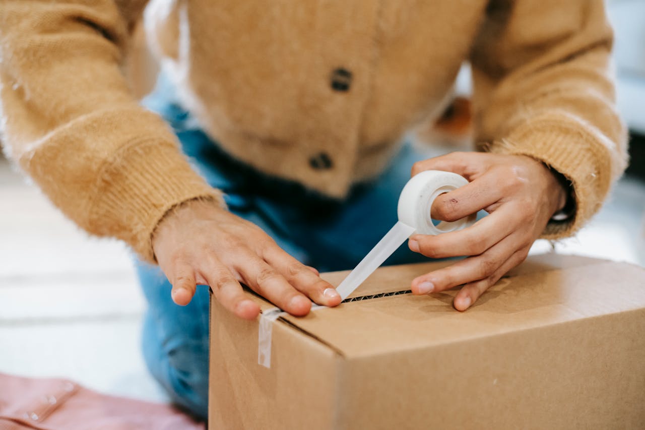 journey A woman uses tape to seal a cardboard box at home, preparing for shipping or moving.