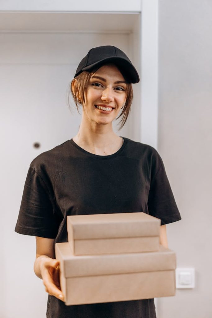 pexels photo 6969839 A cheerful delivery woman holds packages while standing indoors, wearing a black cap and uniform.